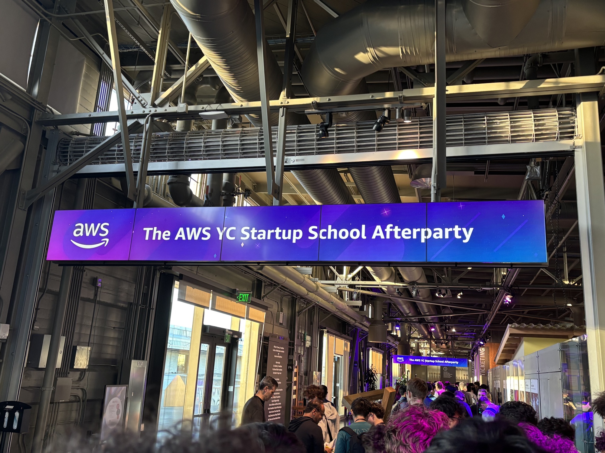 a purple screen reading The AWS YC Startup School Afterparty above a crowded hallway